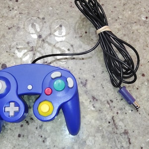 May include: A blue video game controller featuring a central green A button, a red B button, a yellow C-stick, a gray D-pad, two gray shoulder buttons, and a white analog stick, connected to a long black cord with a purple connector plug.