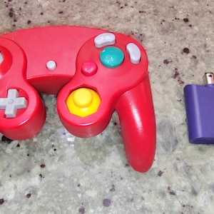 May include: A red video game controller featuring a gray analog stick, a yellow C-stick, a green A button, gray X and Y buttons, and a gray D-pad, accompanied by a small purple rectangular memory card accessory with a connector plug.
