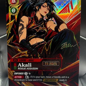 Op de afbeelding: Een holografische verzamelkaart met het personage Akali, getiteld Rogue Assassin, in een rood en zwart kleurenschema. De kaart toont de tekst Akali, Rogue Assassin, T1/2025 en spelinstructies, inclusief een handtekening aan de rechterkant.