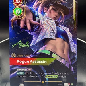 May include: A collectible trading card featuring a character named Akali, titled Rogue Assassin. The card displays a holographic finish, a blue and silver color palette, and game text including Empower 3 and an action description. The character wears a visor and a metallic jacket, with the name Akali written in green script.