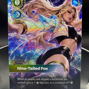 May include: A holographic trading card featuring a long-haired character with fox ears, wearing a black top and shorts. The card displays the text Ahri, Nine-Tailed Fox, and game mechanics. The background has a blue, crystalline, reflective pattern.
