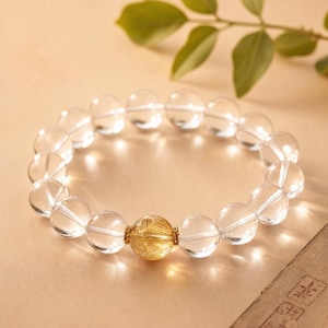 May include: A beaded bracelet composed of smooth, transparent clear quartz spheres and a single central golden rutilated quartz bead. The central bead is flanked by two small, textured gold-toned metal spacers, creating a circular arrangement.