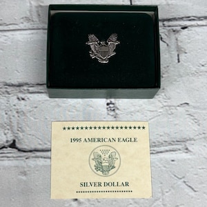 May include: A silver American Eagle emblem pin inside a dark green box with a dark green velvet interior. A card below the box reads "1995 AMERICAN EAGLE SILVER DOLLAR".