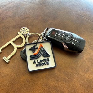 May include: A metal keychain set featuring a gold-toned letter R charm, a small circular charm, and a square white rubberized tag with a black and orange mountain graphic and the text A LAYER ABOVE, attached to a black automotive key fob.