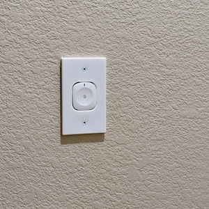 May include: A white rectangular wall-mounted smart home controller featuring a central square button with a circular G logo and a small vertical indicator light at the top, secured to the wall with two visible screws at the top and bottom.