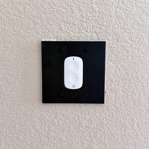 May include: A white smart home button with two circular buttons and a small indicator light, featuring a Google logo at the bottom. The device is mounted on a square black wall plate secured with four black screws against a textured wall.