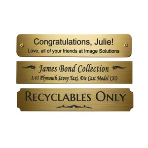 May include: Three gold-colored metal plaques with black text. The top plaque reads "Congratulations, Julie! Love, all of your friends at Image Solutions". The middle plaque reads "* James Bond Collection * 1:43 Plymouth Savoy Taxi, Die Cast Model IXO". The bottom plaque reads "RECYCLABLES ONLY".