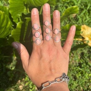 May include: A collection of eight silver rings, each featuring a smooth, oval-shaped pink stone. The rings are worn across four fingers, with two rings on each finger. A silver chain-link bracelet with a small charm is worn on the wrist.