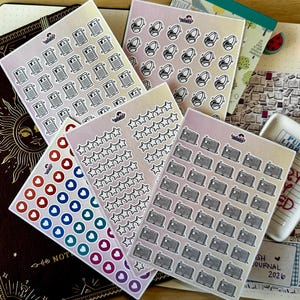 Könnte beinhalten: Eine Sammlung von Aufkleberbögen mit verschiedenen Symbolen wie Büchern, Kopfhörern, Sternen und Herzen, angeordnet auf einem dunkelbraunen Notizbuch mit goldenem Sonnendesign und einer weißen Keramikschale mit der Aufschrift Public Library und Library Card.