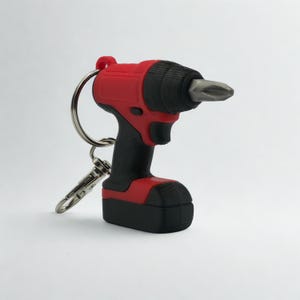 Puede incluir: Un llavero en forma de taladro en miniatura con cuerpo rojo y negro y una punta de destornillador de metal plateado en la parte delantera. La herramienta está unida a una anilla de metal plateado y a un pequeño cierre de mosquetón.