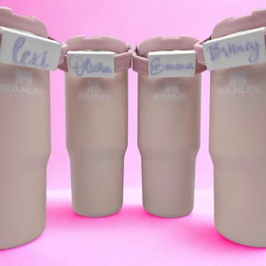 May include: Four light pink Stanley tumblers arranged in a row, each featuring a white rectangular name tag attached to the handle. The tags display the names Lexi, Olivia, Emma, and Britney in a purple cursive script font on a white background.