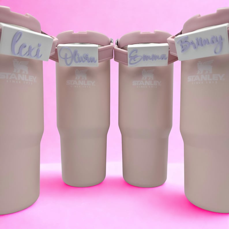 May include: Four light pink Stanley tumblers arranged in a row, each featuring a white rectangular name tag attached to the handle. The tags display the names Lexi, Olivia, Emma, and Britney in a purple cursive script font on a white background.