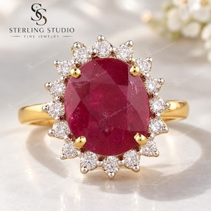 May include: A gold ring featuring a large oval-cut deep red ruby centre stone surrounded by a halo of small round brilliant-cut white diamonds. The ruby is held by four gold prongs, and the diamonds are set in a scalloped, sunburst-style frame.