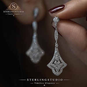 Puede incluir: Un par de pendientes colgantes de tono plateado con intrincado trabajo de filigrana metálica y piedras transparentes incrustadas. El diseño incluye una parte superior puntiaguda conectada a una base en forma de abanico. El texto STERLINGSTUDIO Timeless Elegance es visible.