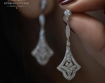 May include: A pair of silver-toned drop earrings featuring intricate filigree metalwork and embedded clear stones. The design includes a pointed top section connected to a larger, fan-shaped base. The text STERLINGSTUDIO Timeless Elegance is visible.