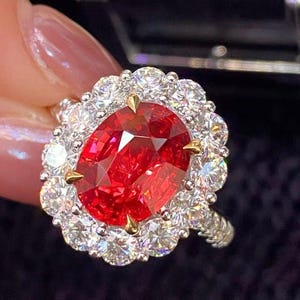 May include: A ring featuring a large, oval-cut vibrant red gemstone set in yellow gold prongs, surrounded by a halo of numerous round, brilliant-cut clear white diamonds set in a silver-toned metal band with a textured, pavé-style shank.