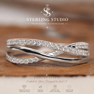 Puede incluir: Un anillo cruzado de plata con dos bandas entrelazadas, una pulida y lisa, y la otra incrustada con pequeñas piedras redondas transparentes. El anillo se presenta con el texto de marca STERLING STUDIO y varios iconos.