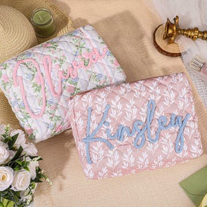 Peut inclure: Deux pochettes rectangulaires matelassées avec fermetures éclair. L'une présente un fond blanc avec un motif floral rose et bleu et le prénom Olivia en écriture rose. L'autre est rose poudré avec un motif de feuilles blanches et le prénom Kinsley en bleu.