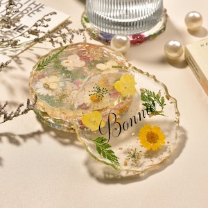 May include: A set of round, clear resin coasters with wavy edges, embedded with various dried flowers, green leaves, and gold flakes. One coaster features the name Bonnie in black cursive script. The coasters have a glossy, smooth finish.