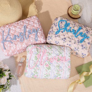 May include: Three rectangular quilted cosmetic bags featuring floral patterns and personalized names in raised script lettering. One bag is pink with white leaves and the name Kingsley, one is beige with blue flowers and the name Shirley, and one is striped with pink flowers and the name Olivia.