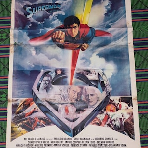 Puede incluir: Un póster vintage de la película Superman con una ilustración central de Superman volando hacia adelante con una capa roja. Debajo, un gran escudo S metálico contiene retratos del reparto, con créditos de producción y nombres al pie.