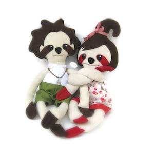 May include: Two plush sloth toys, one wearing green shorts and a necklace, the other wearing a red and white cherry print dress, hugging each other.
