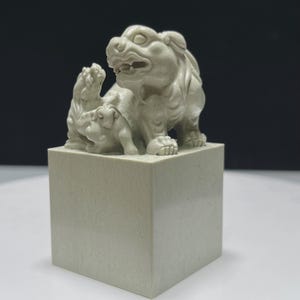 May include: A light gray, square-based seal featuring a carved sculpture of a large mythical creature with an open mouth standing beside a smaller creature. The base is a smooth, solid cube with a matte finish, supporting the detailed animal figures.
