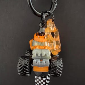 May include: A toy monster truck keychain with black wheels, an orange body, and a clear orange accent piece. The keychain has a black metal clasp and a checkered flag design.