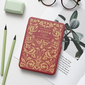 May include: A red book cover featuring gold embossed floral patterns and the text "JANE AUSTEN EMMA ILLUSTRÉ PAR HUGH-THOMSON". The cover has a textured surface and is positioned on a white background with two light green pencils and a plant illustration.