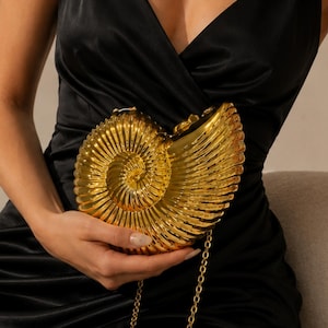 May include: A gold-toned, nautilus shell-shaped clutch bag with a metallic finish and a textured, ribbed surface. The clutch features a long, thin gold-coloured chain strap and is held against a black satin slip dress with thin spaghetti straps.