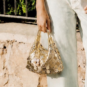 May include: A metallic gold handbag featuring a circular sequin pattern and a structured handle. The bag is held at the side, showcasing its reflective surface and small dangling charms attached to the base of the handle hardware.