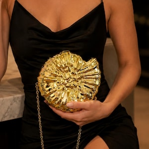 May include: A metallic gold clutch bag shaped like a spiraled ammonite fossil. The handbag features a textured, ridged surface with a high-shine finish and includes a thin gold chain strap for carrying. The design is circular and compact.