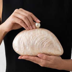 May include: A shell-shaped clutch bag with a glossy, pearlescent cream finish and a smooth texture. The bag features a single, round, white pearl accent at the top centre, serving as a clasp mechanism for the curved, rigid, clam-like structure.