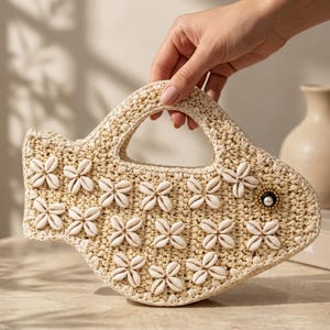 Pode incluir: Uma bolsa em forma de peixe feita de material trançado bege, com uma alça recortada no topo. O exterior é decorado com várias conchas de búzios brancas dispostas em padrões florais e um olho feito de uma conta preta.