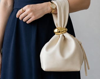 May include: A cream-coloured pouch handbag featuring a gathered top closure secured by a polished gold-tone metal ring. The bag has a soft, smooth texture and includes a matching cream-coloured strap that loops through the gold ring hardware.