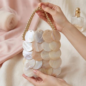 Puede incluir: Un bolso pequeño construido con discos circulares de nácar superpuestos en tonos crema, beige y blanco iridiscente. El bolso cuenta con un asa superior única hecha de una hebra continua de cuentas de metal pulido en tono dorado.