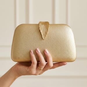 May include: A gold-coloured clutch bag with a textured, metallic finish and a prominent, leaf-shaped clasp centred at the top. The rectangular bag features rounded corners and a solid, uniform appearance, showcasing a distinct, detailed leaf design.