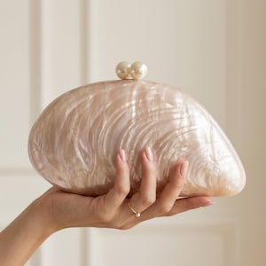 Pode incluir: Uma bolsa clutch semicircular com um acabamento perolado brilhante que imita a aparência de uma concha. A bolsa apresenta um fecho com dois detalhes de pérolas brancas redondas, proporcionando uma textura de superfície lisa e iridescente.