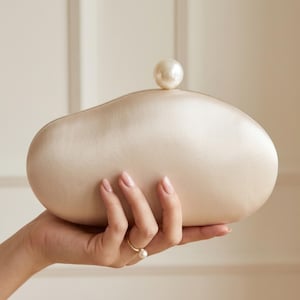 May include: A smooth, cream-coloured satin clutch bag with an asymmetrical, rounded shape. The top of the bag features a single, large, spherical white pearl as a clasp closure. The surface has a soft, subtle sheen characteristic of satin fabric.