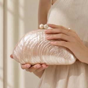 Peut inclure: Une pochette semi-circulaire avec une finition nacrée semblable à un coquillage dans des tons crème et rose pâle. Le sac présente une fermeture à fermoir surmontée de deux sphères rondes blanches rappelant des perles. La surface est texturée.