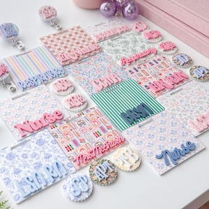 May include: A collection of square badge reels and circular badge reels featuring various patterns including floral, striped, and medical icons. The badge reels display raised lettering in pink, blue, and white, with text such as Nurse, ER Nurse, and Bryn.