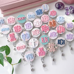 May include: A collection of circular badge reels featuring various designs including pastel stripes, floral patterns, and animal prints. Each reel displays raised lettering or symbols such as monograms, the text love like jesus, or a cross.