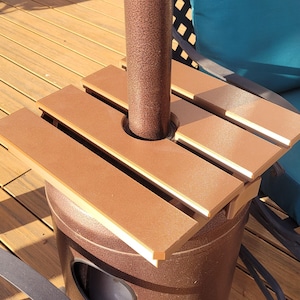 May include: A brown, square, slatted table attachment designed to fit around a central pole. The table features a smooth, matte finish and a circular cutout in the center to accommodate the pole, providing a flat surface for items on a patio.