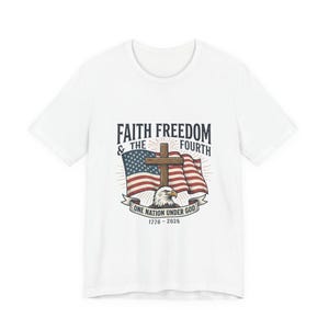 Könnte beinhalten: Ein weißes T-Shirt mit einem Grafikdruck, der eine amerikanische Flagge, ein Holzkreuz und einen Weißkopfseeadler zeigt. Der Text lautet FAITH FREEDOM & THE FOURTH, ONE NATION UNDER GOD und 1776 - 2026. Das Motiv ist mittig auf der Brust platziert.
