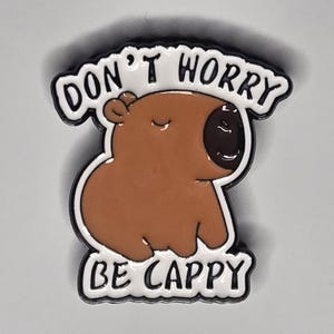 Puede incluir: Un pin de esmalte marrón que presenta un capibara de dibujos animados con los ojos cerrados. El pin tiene un borde blanco con texto negro que dice DON'T WORRY en la parte superior y BE CAPPY en la parte inferior. Tiene una nariz marrón.