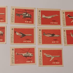 Pode incluir: Uma coleção de dez etiquetas retangulares de caixas de fósforos com fundos vermelhos e ilustrações pretas de várias aeronaves e helicópteros soviéticos. Cada etiqueta exibe um nome de modelo em cirílico, incluindo IL-62, KA-26, TV-144, YK-40, MI-10, TV-134, AN-12, BE-30, MI-8 e TV-154, dispostos em uma grade.