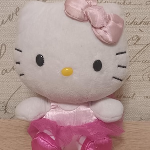 Puede incluir: Un peluche de Hello Kitty que representa un personaje de gato blanco con nariz amarilla, ojos negros y tres bigotes a cada lado. El personaje lleva un lazo rosa claro brillante, un corpiño de satén a juego, un tutú de tul rosa oscuro.