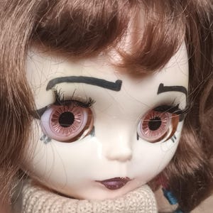 Könnte beinhalten: Ein Puppenkopf mit welligem braunem Haar, großen rosa Augen mit detaillierten Iris und langen schwarzen Wimpern. Das Gesicht hat aufgemalte schwarze Augenbrauen, dunkle Lippen und einen beigen Strickkragen, der unten im Bild zu sehen ist.