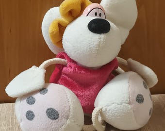 May include: A plush toy featuring a white mouse with large ears, a black nose, and pink eyelids. The mouse wears a bright pink shirt and a yellow bow on its head. Its large white feet are decorated with three grey circular patches on each sole.