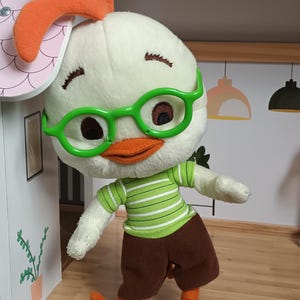 Può includere: Un peluche di Chicken Little, con testa bianca, occhiali verdi, becco e cresta arancioni e maglietta a righe verdi e bianche. Il giocattolo indossa pantaloncini marroni e piedi arancioni.